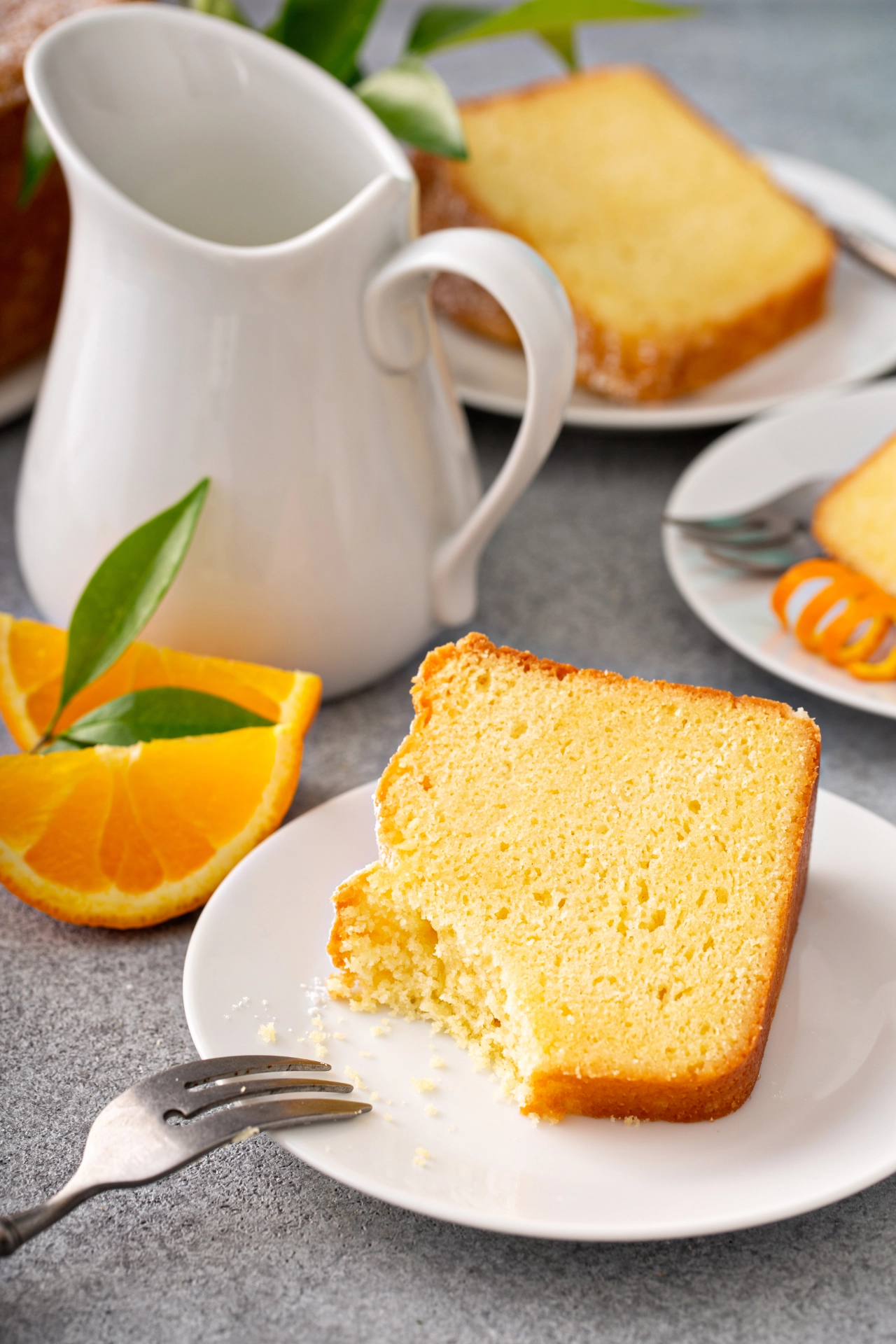 Rebanada de bizcocho esponjoso de naranja en un plato blanco, con gajos de naranja y jarra de cerámica al fondo