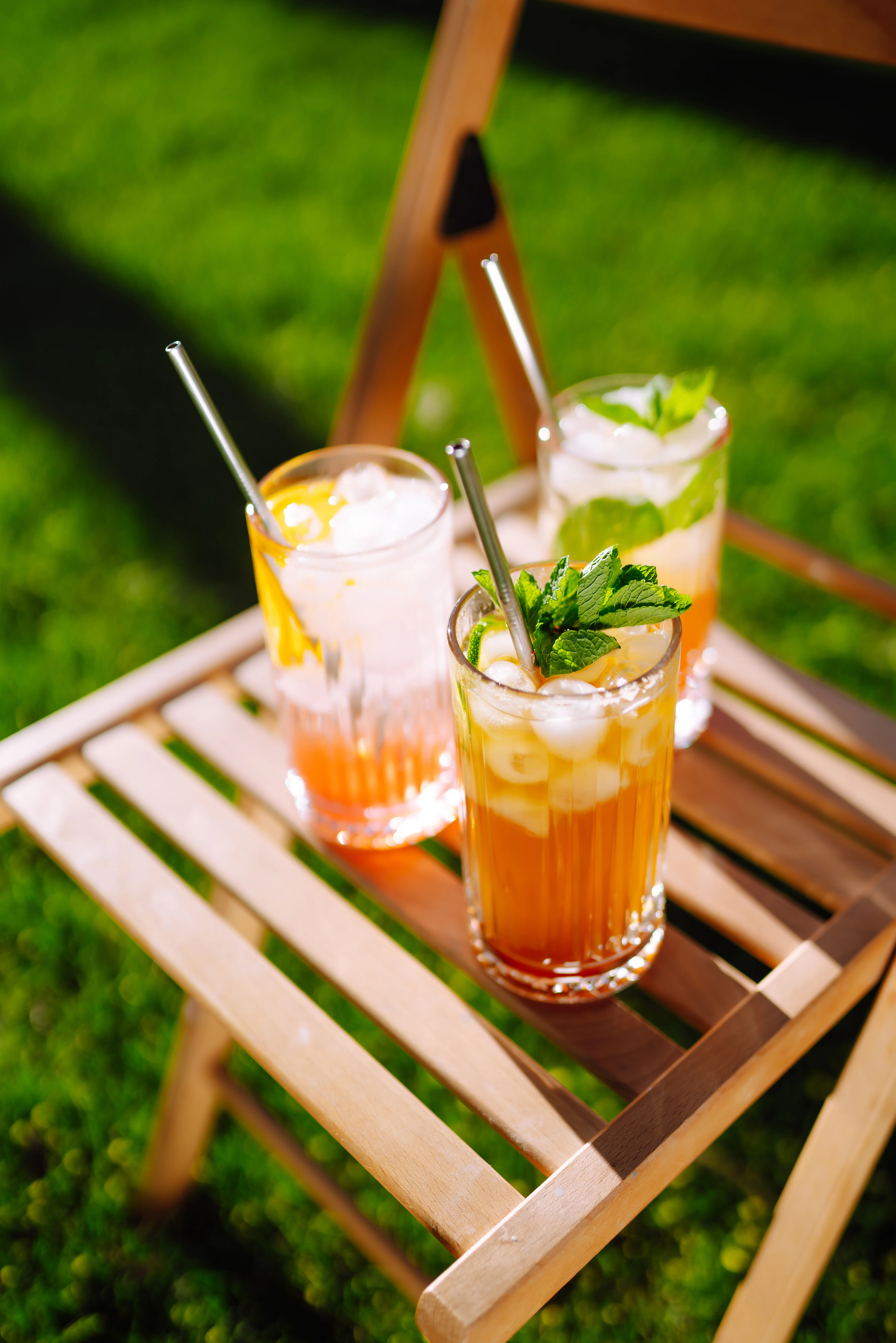 Tres vasos altos con limonada helada, hielo y hojas de hierbabuena, sobre silla de madera y césped