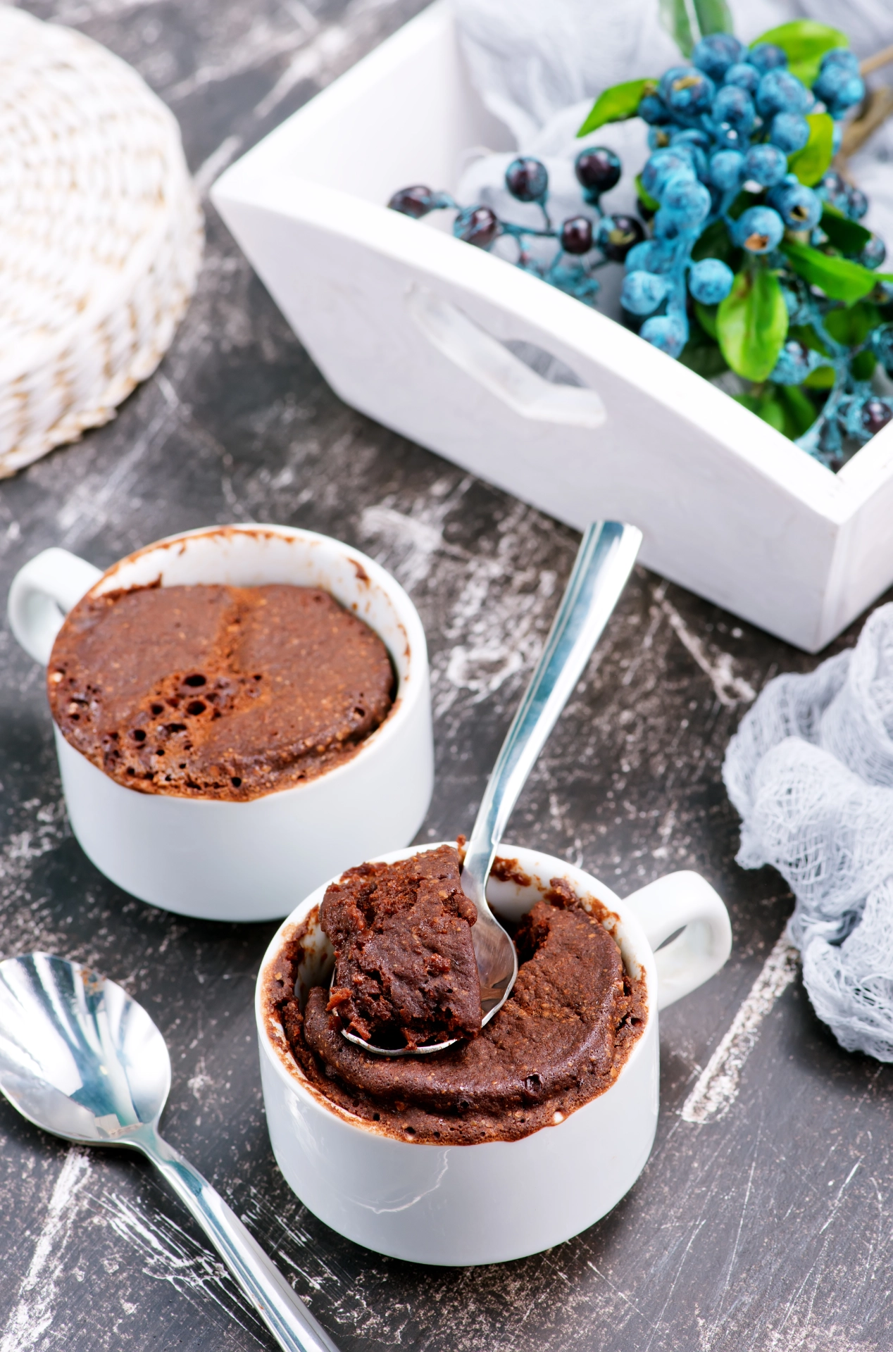 Dos tazas blancas con mugcake de chocolate, una abierta con cuchara, sobre superficie oscura