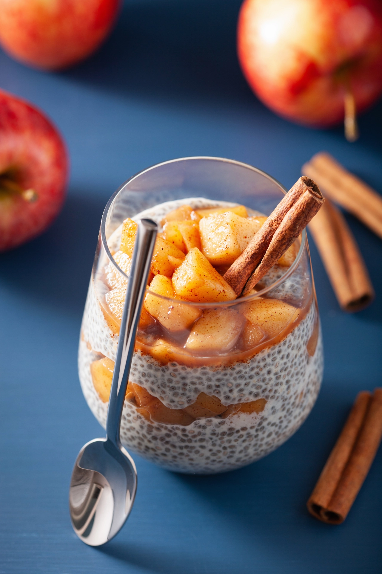 Copa con yogur en capas, cubos de manzana en caramelo y rama de canela, manzanas rojas al fondo sobre superficie azul